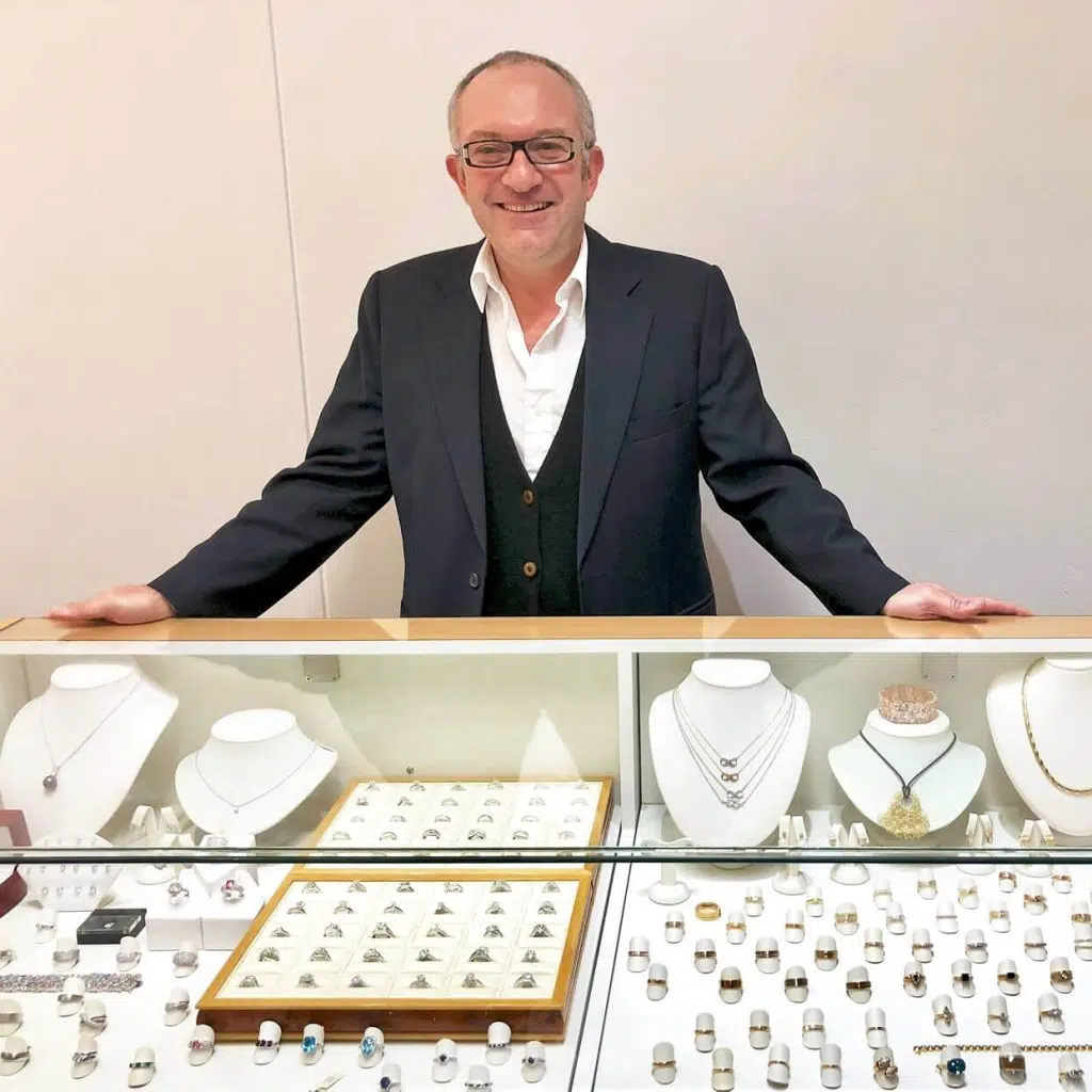 Photo of Mark Nathan the jewellery designer standing next to a display which features rings, diamonds, necklaces and pendants